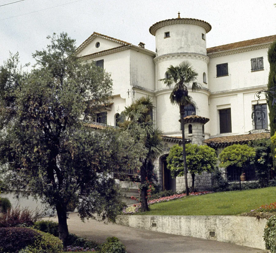 Una villa blanca con una torre cilíndrica en la esquina y tejados de tejas tras un olivo, palmeras y un césped recortado, fotografiada en Grasse.