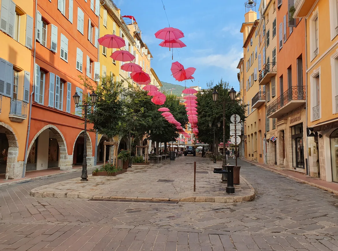 Una callejuela empedrada del Grasse antiguo flanqueada por casas con soportales naranjas y amarillas, con paraguas rosas colgados cruzando el cielo.