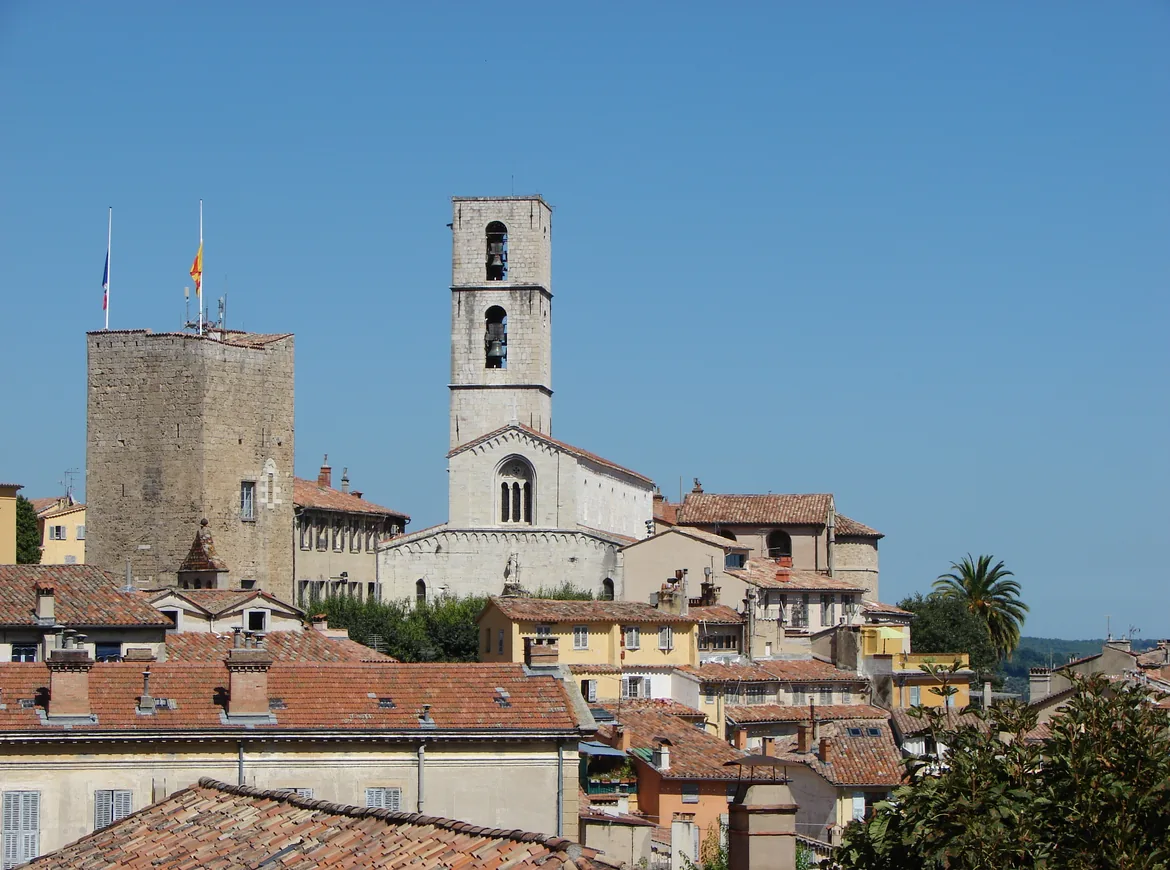 El casco antiguo de Grasse: una torre campanario de piedra y una torre del homenaje cuadrada sobre tejados de terracota y casas ocre bajo un cielo azul despejado.