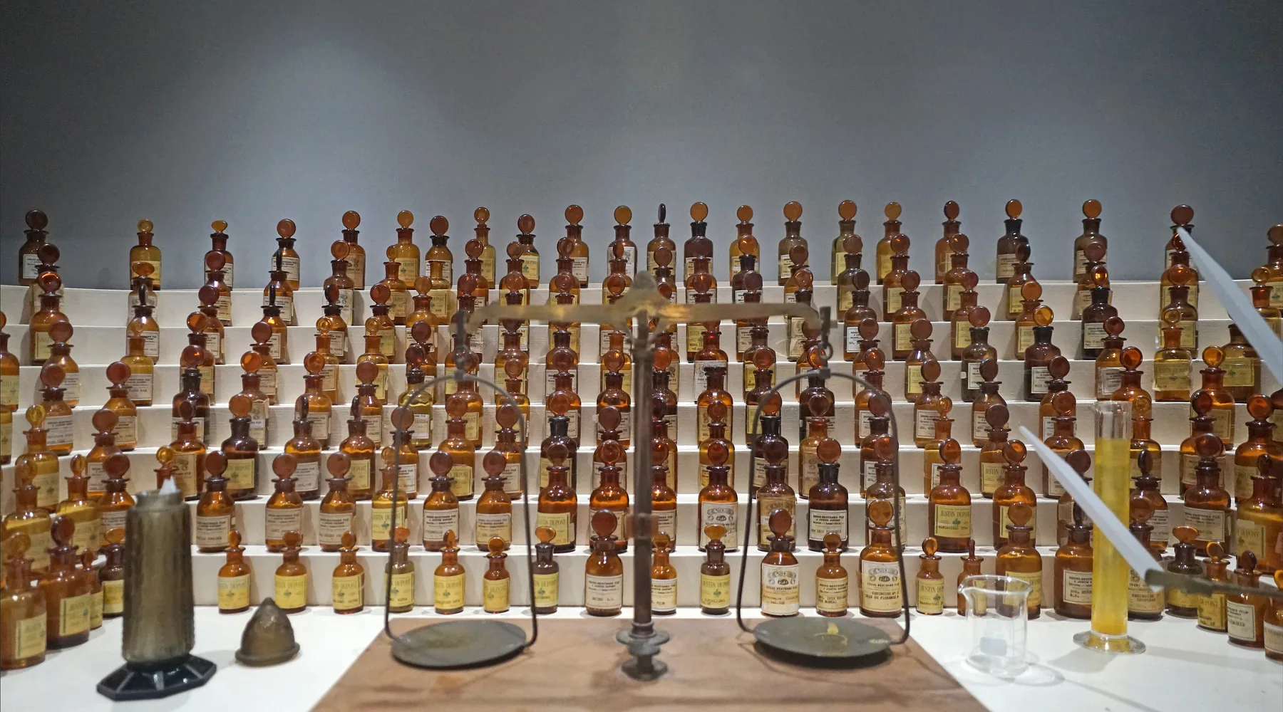 Un órgano de perfumista escalonado con frascos de vidrio ámbar tras una balanza de latón y una probeta graduada, en un museo de Grasse.