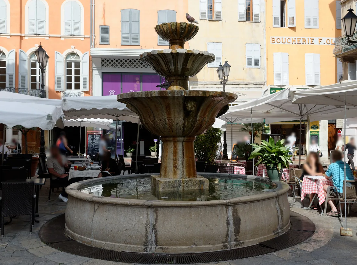 Una fuente de piedra de tres niveles en una plaza de Grasse, con mesas de café bajo sombrillas blancas y casas señoriales de color melocotón al fondo.